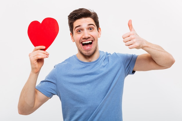 Cheerful man holding paper heart and showing thumb up