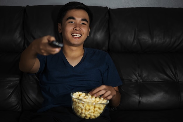 Cheerful man holding remote control and watching TV while sitting on sofa at night