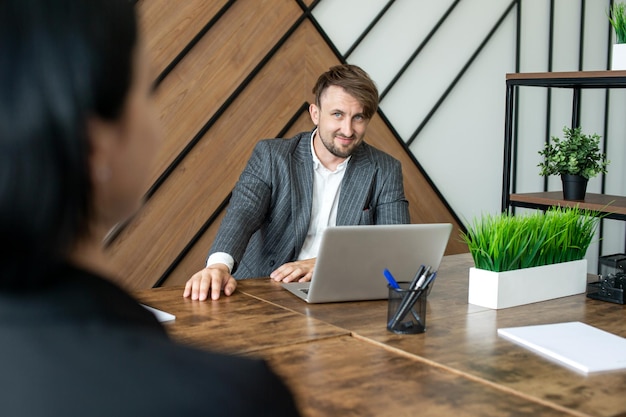 A cheerful man in a jacket is sitting at his desk in the office with a laptop