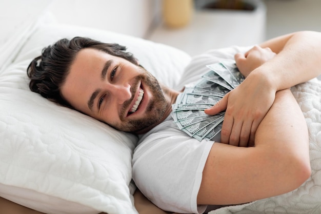 Cheerful man laying in bed hugging cash