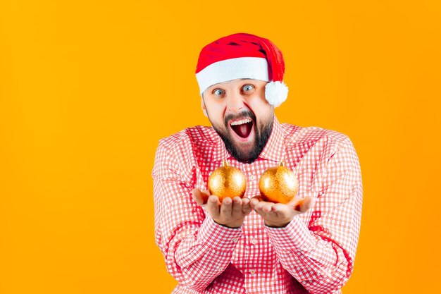 クリスマスの黄色の背景にサンタクロースの帽子をかぶった陽気な男が彼の前にクリスマスツリーの黄金色のボールを保持します-おもちゃ