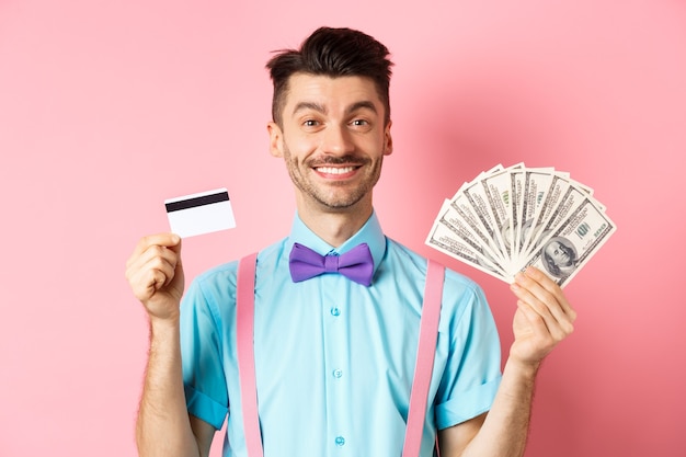 彼のお金とプラスチックのクレジットカードを見せて、カメラに幸せそうに笑って、ピンクの背景の上に立っている陽気な男。