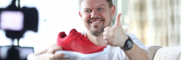 Cheerful man sit on couch, hold red sneaker in his hand and show thumbs up. Blogger shoot video at home.