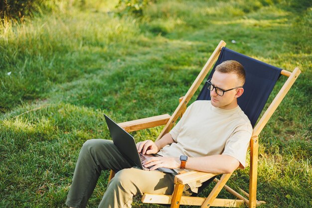 陽気な男性が自然の中で椅子に座り、ラップトップでオンラインで仕事をする 男性が旅行し、ラップトップ コンピューターでリモートで仕事をする 自然の中でオフィスで仕事をする