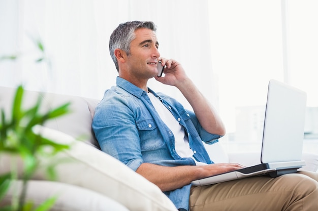 Cheerful man sitting on couch phoning and using laptop