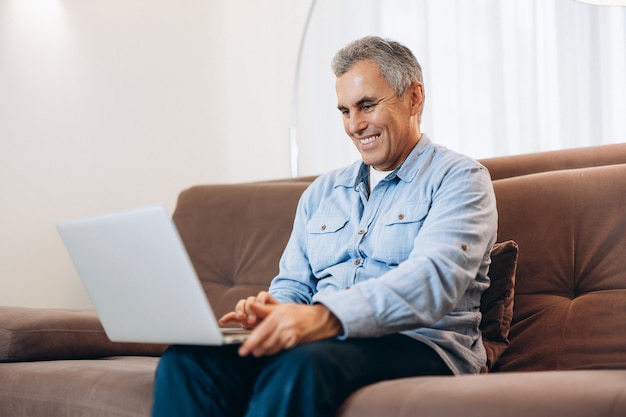 Cheerful man sitting on sofa, using laptop at home in cozy living room