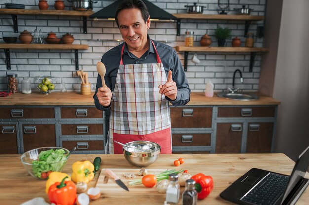 Cheerful man stand at table in kitchen and look on camera. He hold wooden spoon and show big thumb up. Laptop and colorful vegetables on table.