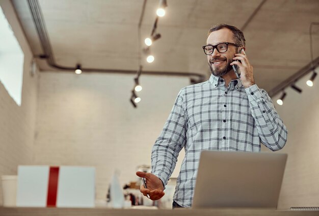 Cheerful man talking on cellphone and using notebook in cafe