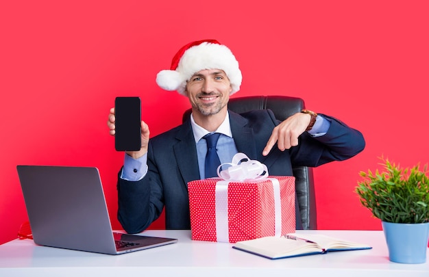 Foto l'uomo allegro indossa il cappello rosso di babbo natale e gli occhiali da festa tengono la confezione regalo che presenta lo smartphone