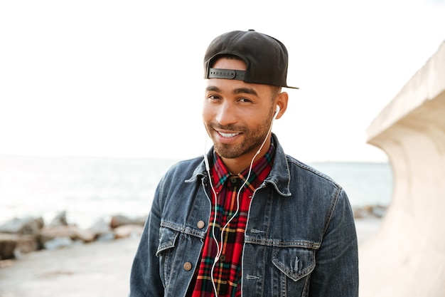 Cheerful man wearing cap walking on the beach while listening music with earphones. Look at camera.