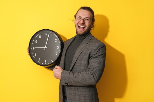 Cheerful man wearing glasses holding big clock on yellow background