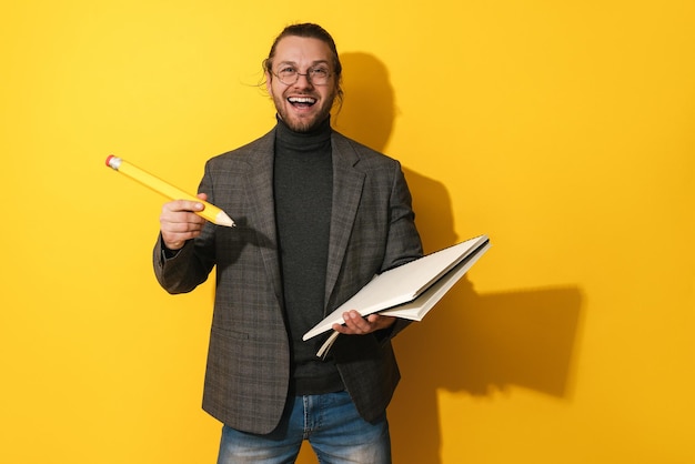 Foto uomo allegro con gli occhiali che tiene grande matita e taccuino su sfondo giallo