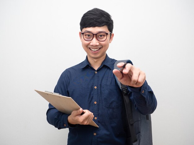 Cheerful man wearing glasses holding clipboard with backpak point pen at you