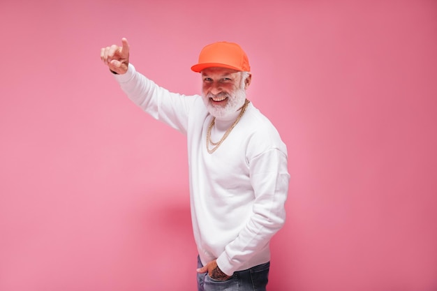 Cheerful man in white sweater and orange cap pointing up on pink background Charming guy with gre beard in light outfit with gold chain posing