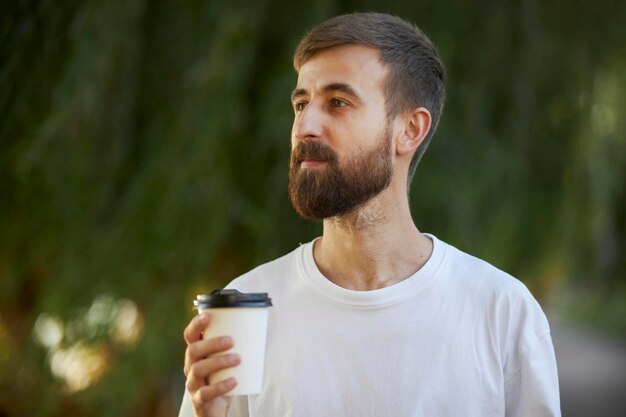 A cheerful man in a white T-shirt drinks coffee from a white paper cup. A white paper cup