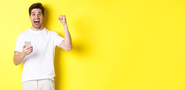 Cheerful man winning on smartphone raising hand up and holding mobile achieve app goal standing over