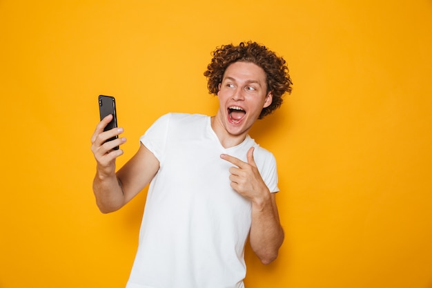 Cheerful man with curly hair smiling and pointing finger at smartphone in hand