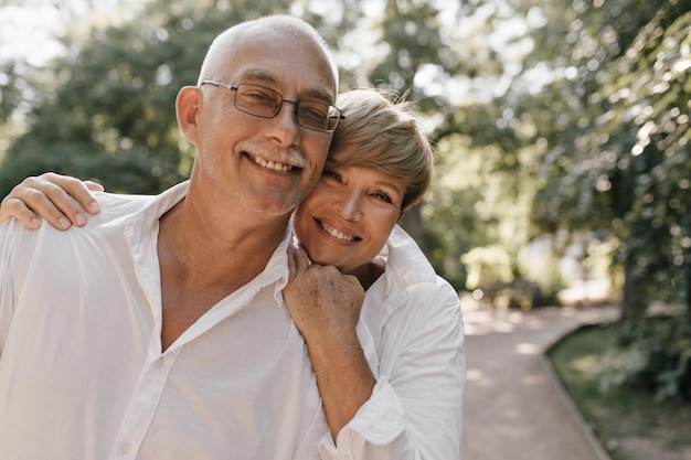 Cheerful man with grey mustache in modern shirt and eyeglasses smiling and posing with short haired lady in white blouse outdoor