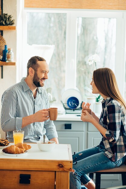 Cheerful man and woman holding mugs while sitting in the kitchen and talking