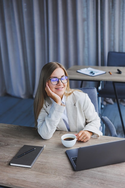 Cheerful manager woman in glasses with laptop smiling at camera during coffee break in office interior happy european woman with caffeine drink posing