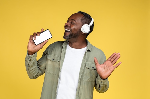 Cheerful mature african american male in casual wireless headphones sings at imaginary microphone and enjoys song