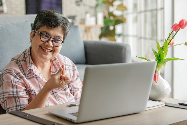 Cheerful mature Asian woman gesticulating and speaking with online colleague while sitting near sofa and working remotely from home in daytime