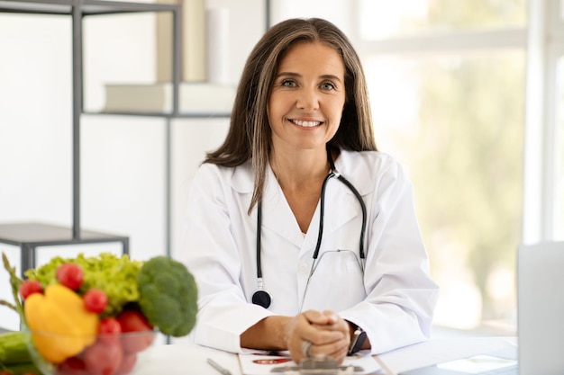 Cheerful mature caucasian woman doctor nutritionist in white coat at table with laptop with organic