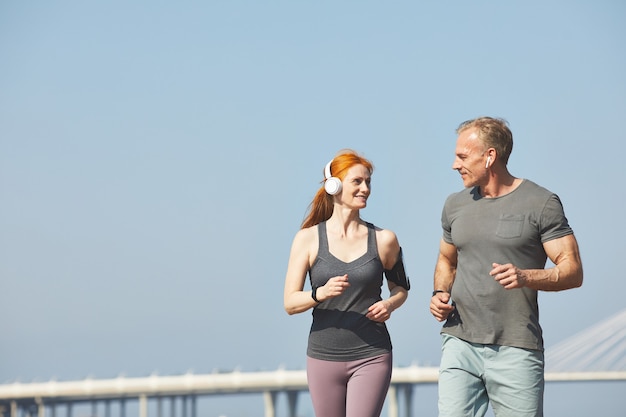 Cheerful mature couple chatting while jogging together over city street, healthy lifestyle concept