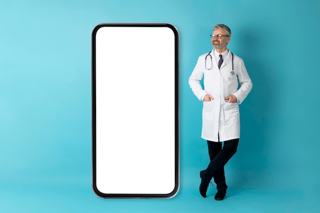 Веселый зрелый врач позирует на смартфоне с макетом на голубом