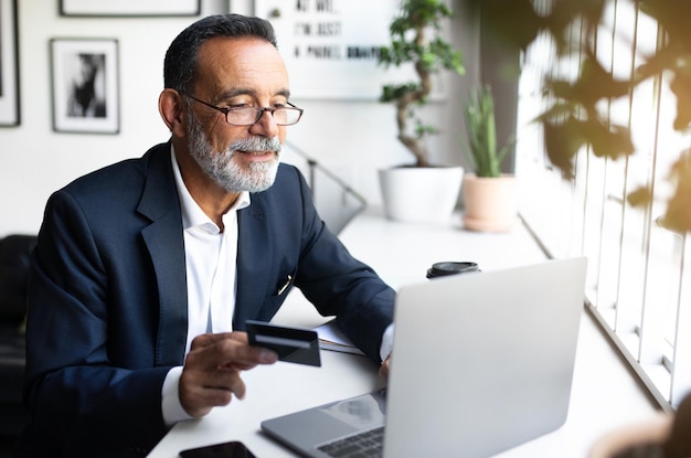 Cheerful mature european man in suit works at laptop uses credit card for investment startup in