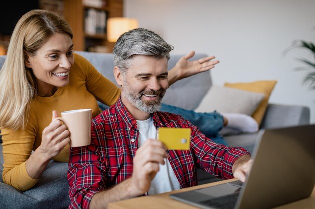 Moglie europea matura allegra con una tazza di bevanda marito con carta di credito e laptop che acquista online