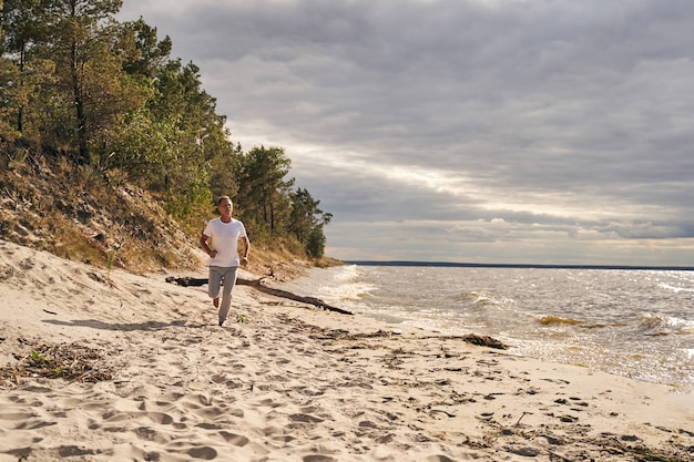 Cheerful mature male is starting day with cardio workout while jogging in solitude along sea shore