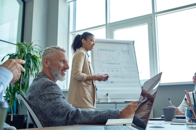 Cheerful mature man sitting at meeting in the modern office and smiling while his young mixed race