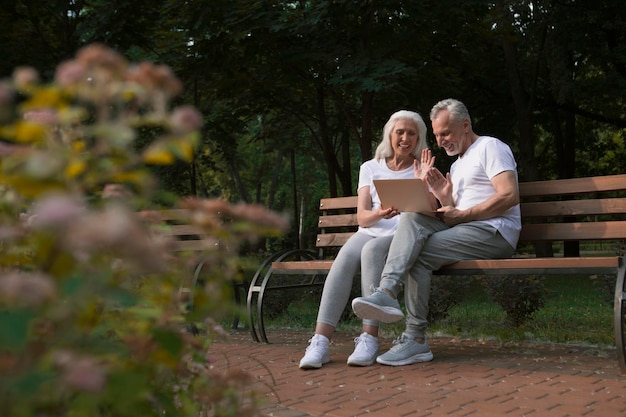 Cheerful mature man and woman with laptop in the park waving to a camera at the video call