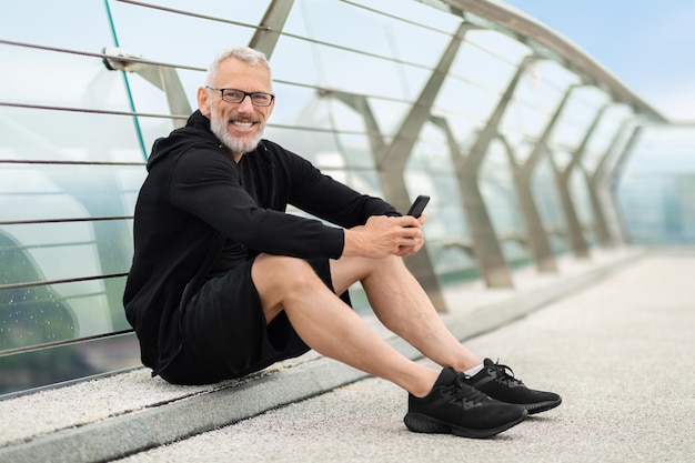 Cheerful mature sportsman having break using smartphone workout outdoor