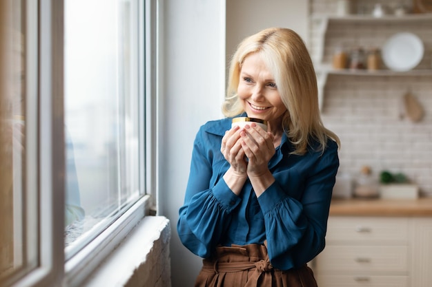 Cheerful mature woman enjoying morning coffee at home standing near window at cozy kitchen holding