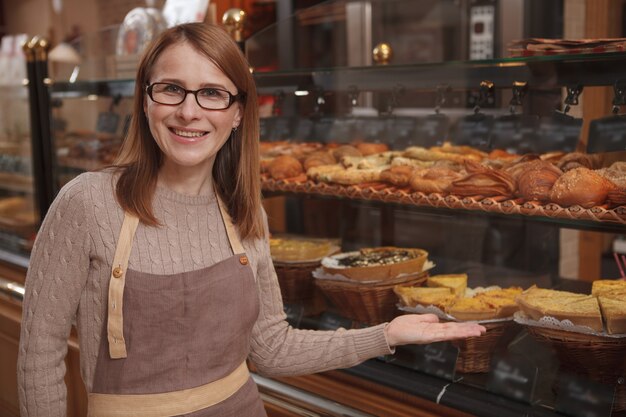 Cheerful mature woman enjoying working at her bakery store, pointing proudly at retail display