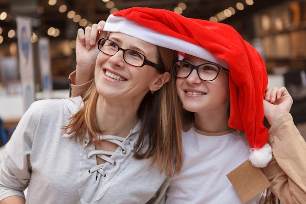 Foto allegra donna matura e sua figlia adolescente ridendo, indossando il cappello di babbo natale