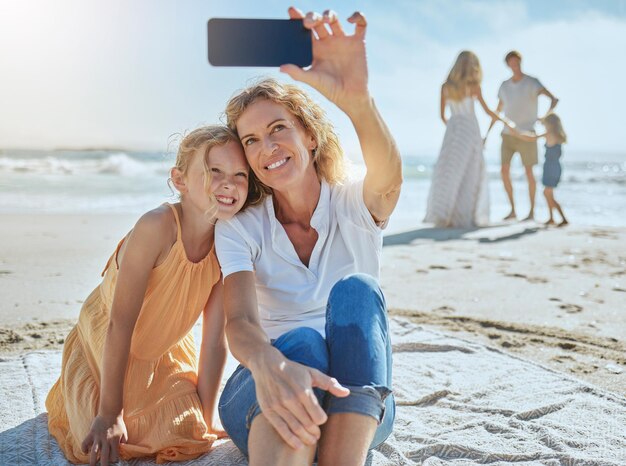 Cheerful mature woman and little girl taking a selfie while sitting on the beach Happy little girl smiling while sitting with her mom or grandmother taking picture on mobile phone while on vacation