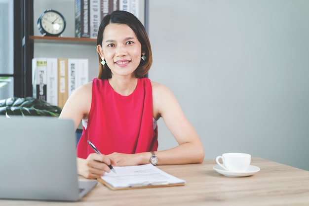 A cheerful middle-aged Asian businesswoman in relax casual dress working at home, checking email on computer laptop, writing on financial accounting document paper. Stock photo