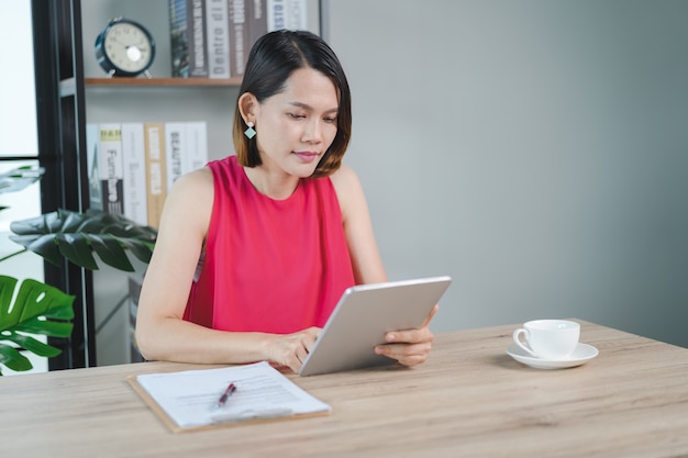 A cheerful middle-aged Asian businesswoman in relaxed casual dress working at home, checking email on computer laptop, writing on financial accounting document paper. Business stock photo