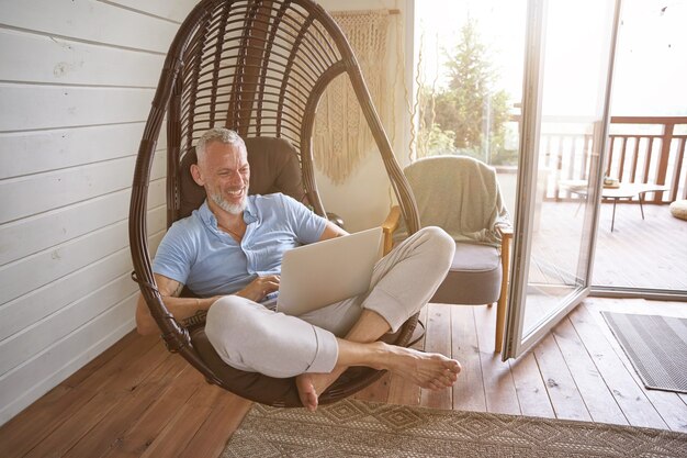 Cheerful middle aged caucasian businessman in casual clothes sitting with laptop on swing chair at