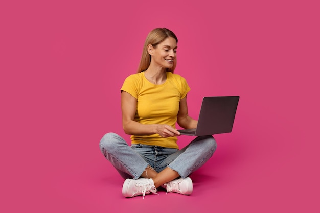 Photo cheerful middle aged european blonde female sitting on floor typing on computer