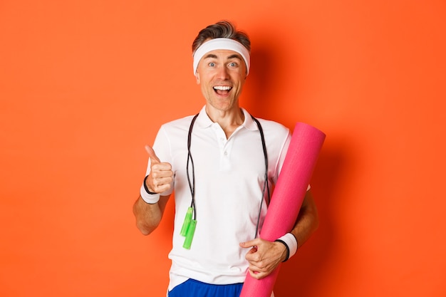 Cheerful middle aged fitness guy, holding skipping rope and yoga mat