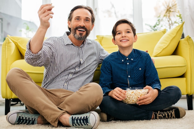 Cheerful middle aged man spending time with his positive son and smiling while enjoying watching TV on the floor at home with popcorn