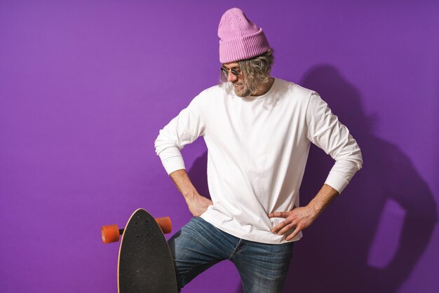 Cheerful middle aged man with longboard wearing white sweatshirt against purple background