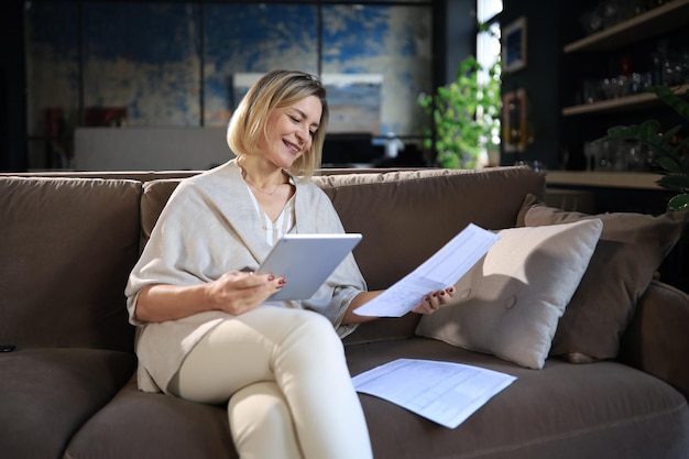 Cheerful middle aged woman sitting on sofa using computer tablet apps looking at screenreading good news in social network shopping or chatting online