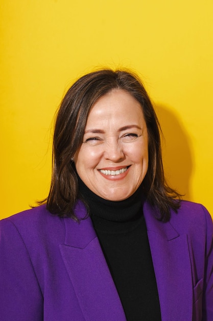 Cheerful middle aged woman wearing purple blazer smiling against yellow background