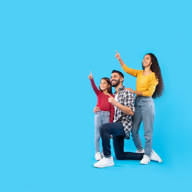 Cheerful middle eastern family pointing fingers upward over blue background