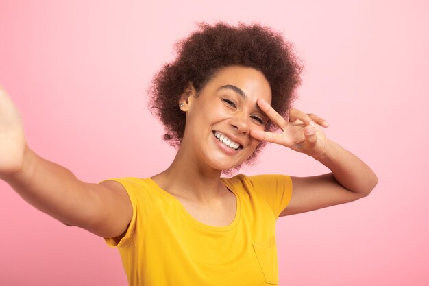 Cheerful millennial african american curly lady student in yellow tshirt making peace sign and selfie